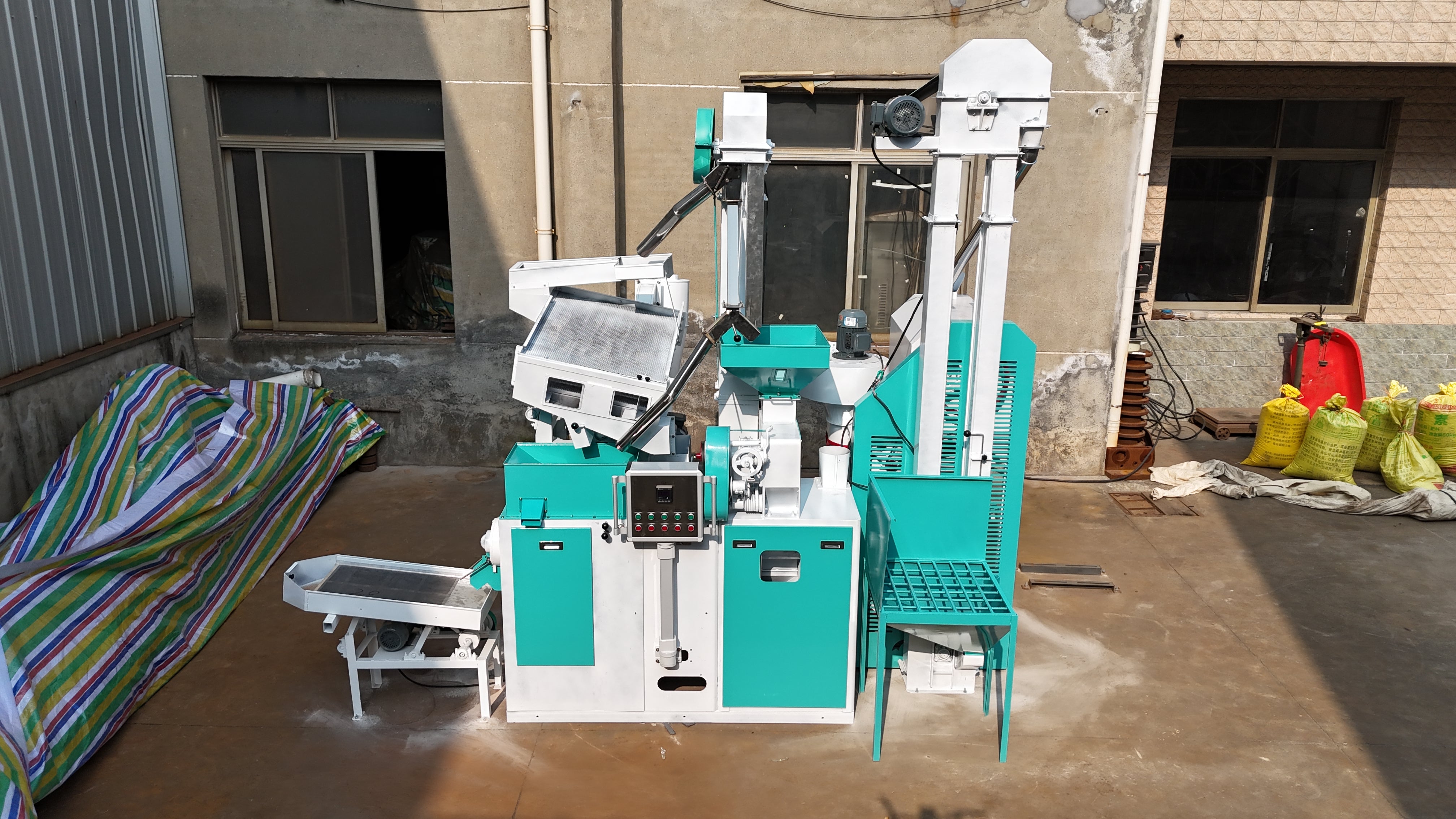 Teal and white rice processing machine in an outdoor setting with a building in the background.